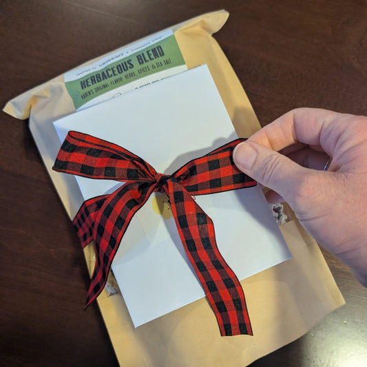 A bow of red-and-black plaid ribbon rests upon a bag of Karen's Herbaceous Blend popcorn.