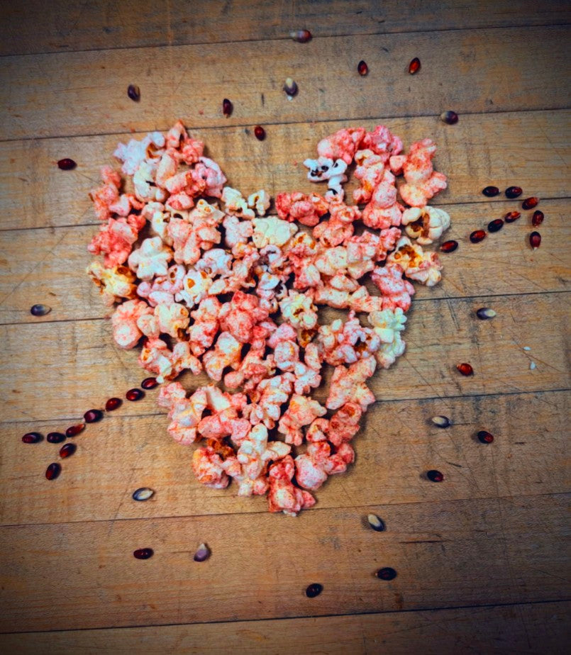 Upon a wooden background, with pink hues, Karen's Candied Berry recipe forms the shape of a heart with red popcorn kernels in the shape of Cupid's arrow "shot" through it.